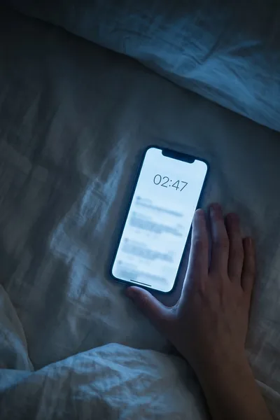 Hand greift nachts zum leuchtenden Smartphone im Bett